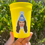 Yellow cup with a sticker of a person wearing a birthday hat and number 25, held in front of green foliage.