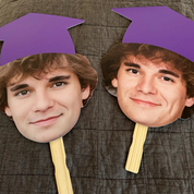 Two cardboard cutouts of a person wearing purple graduation caps on a textured surface.