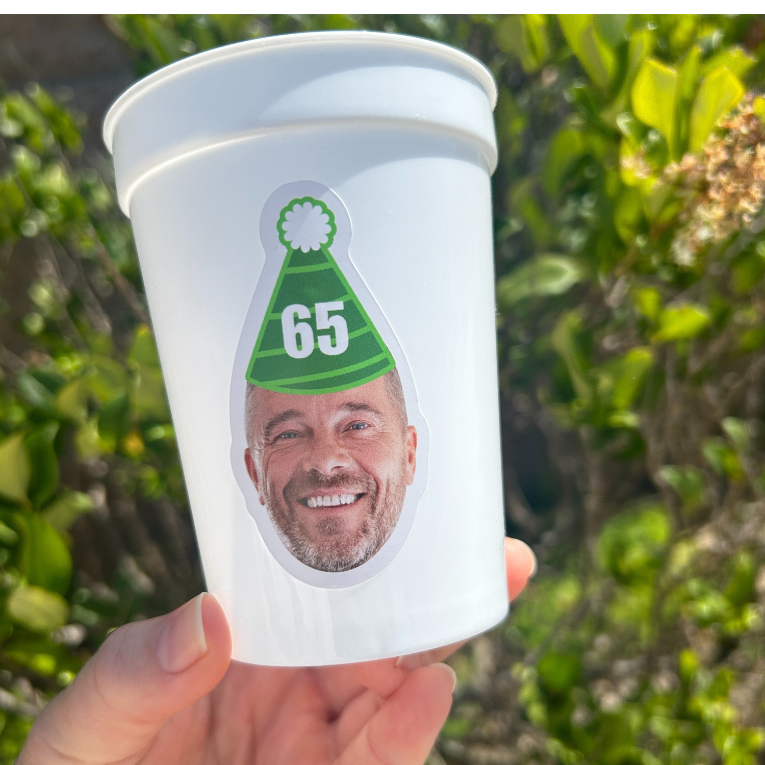 White cup with a printed design of a man wearing a green party hat with the number 65, held in front of green foliage.
