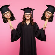Graduate in cap and gown holding cutouts of a face on a pink background