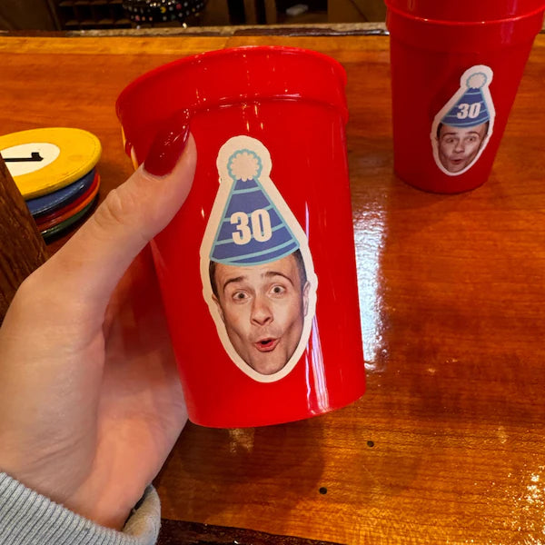 Red cup with a face cutout and '30' hat held by a hand on a wooden table.