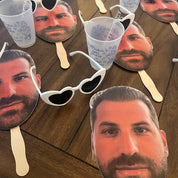 Cardboard cutouts of a man's face with sunglasses and cups on a wooden surface