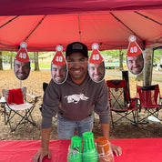Man standing under a red tent with cutout faces and '40' hats, surrounded by camping chairs and a table with drinks.