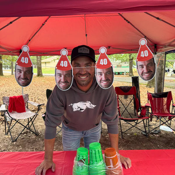 Man standing under a red tent with cutout faces and '40' hats, surrounded by camping chairs and a table with drinks.