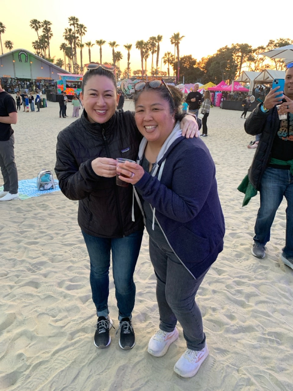 Two women posing together on a beach with palm trees and a sunset in the background.