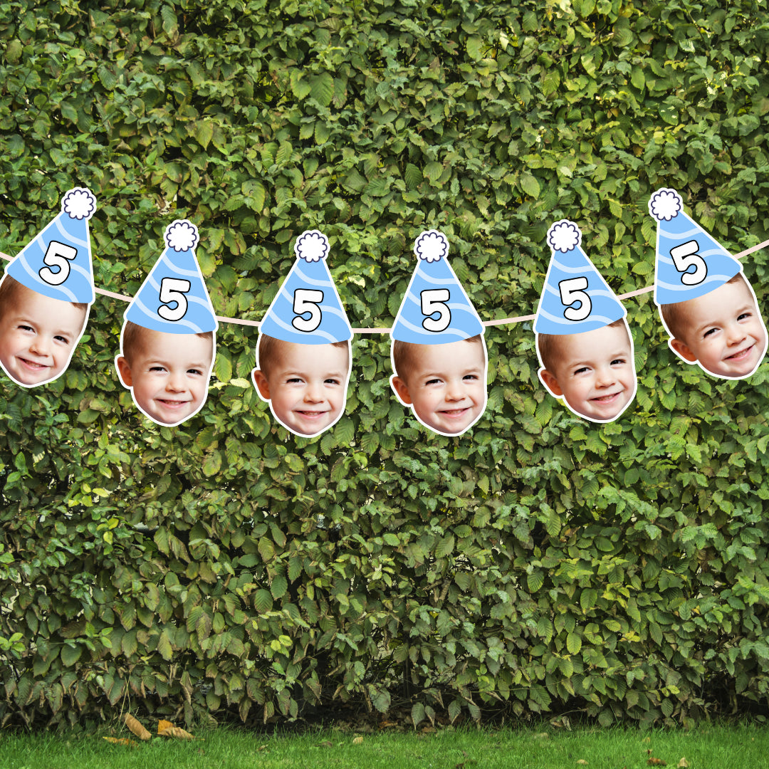 Birthday banner with cut-out faces wearing blue party hats and number 5 on a green hedge background